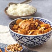 A bowl of massaman curry chicken with a side of rice, and small bowls of white cardamom, cloves and cinnamon sticks