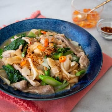 a blue bowl of rad na with chili vinegar and chili flakes on the side