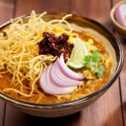 a bowl of khao soi with shallots, lime wedge, cilantro and fried chilies garnish