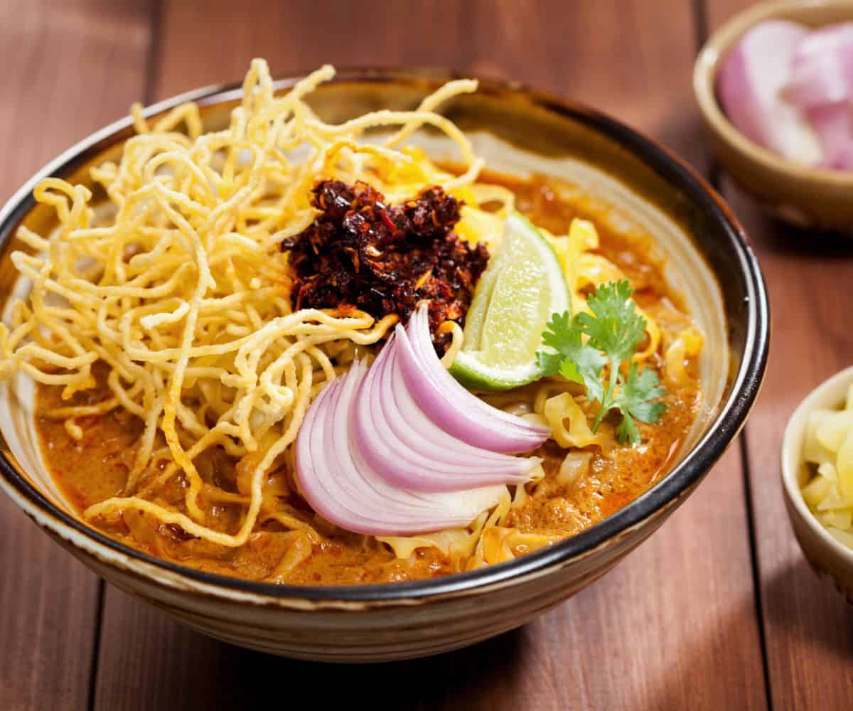 A steaming bowl of kao soi is beautifully topped with crispy noodles, bright and fragran chili crisps, contrasted with the bright red onion slices, lime wedge, and sprig of cilantro.