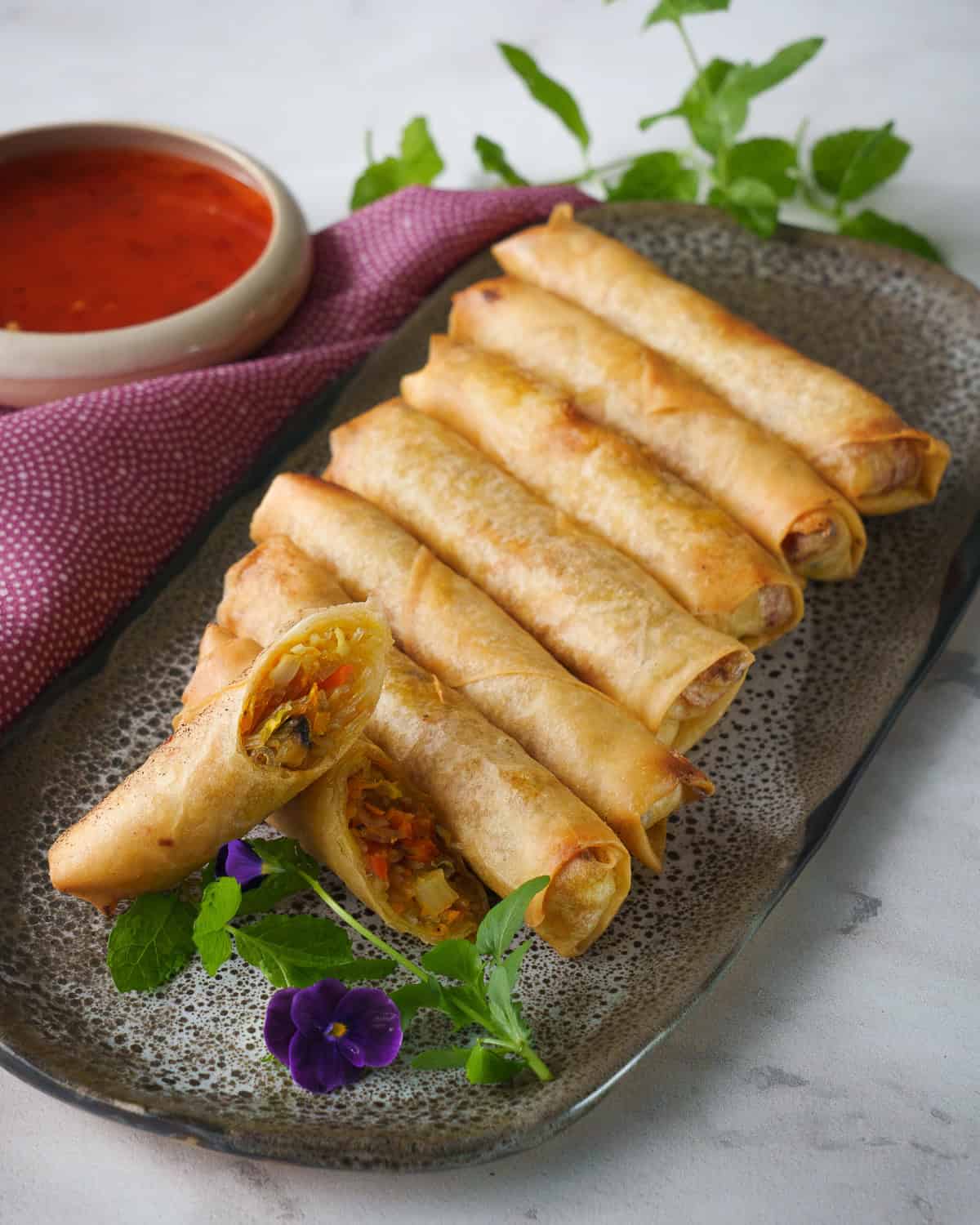 a plate of spring rolls with one cut in half and a bowl of sweet chili sauce on the side and some mint and pansy garnish.