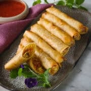 a plate of spring rolls with one cut in half and a bowl of sweet chili sauce on the side and some mint and pansy garnish.