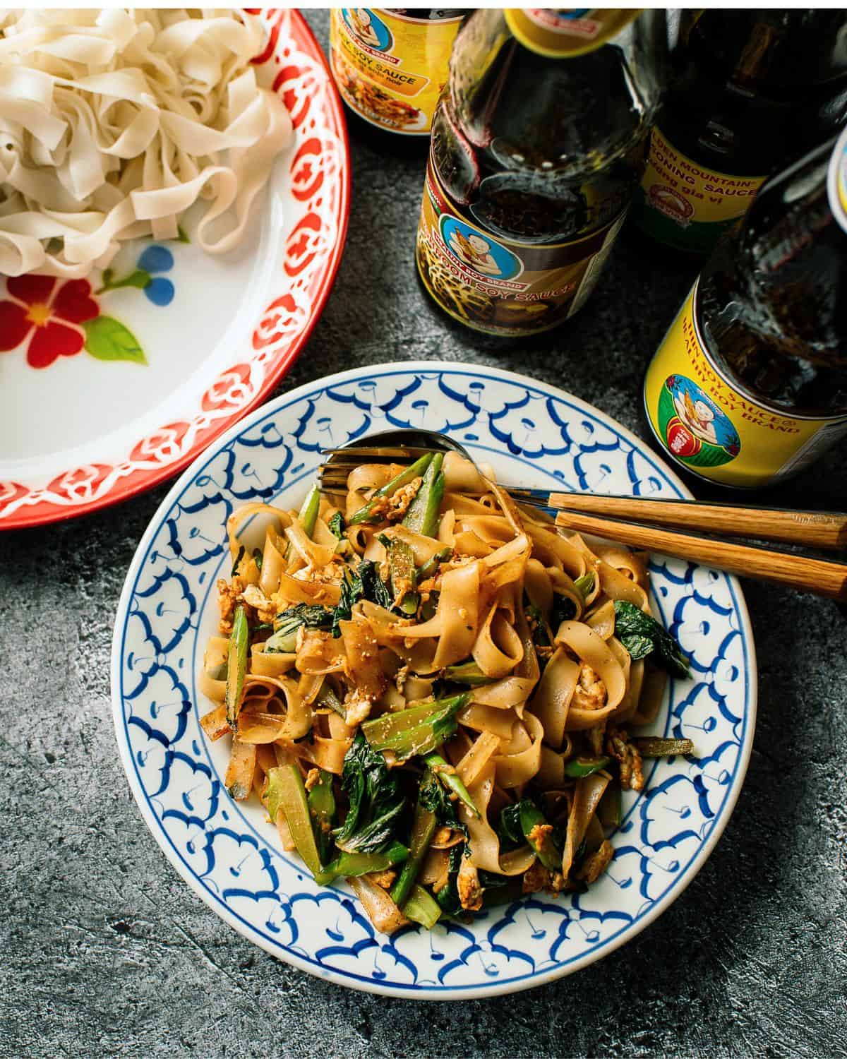 Pad See Ew noodles lay in a georgeous pile on a blue and whote plate surrounded by two bottles of sauce and a red floral white place with wide plain rice noodles. A pair of chopsticks are placed at the top of the blue plate and the background is a grey stone countertop.