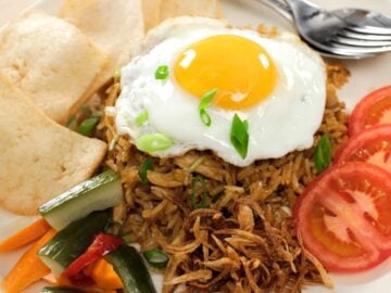 a plate of nasi goreng with a fried egg on top, tomato slices, pickles and shrimp crackers