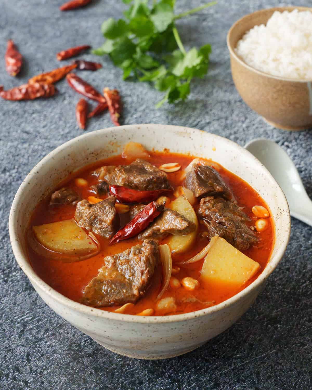 a bowl of massaman curry beef with a bowl of rice on the side with chilies and cilantro in the back.