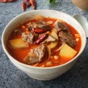 a bowl of massaman curry beef with a bowl of rice on the side with chilies and cilantro in the back.