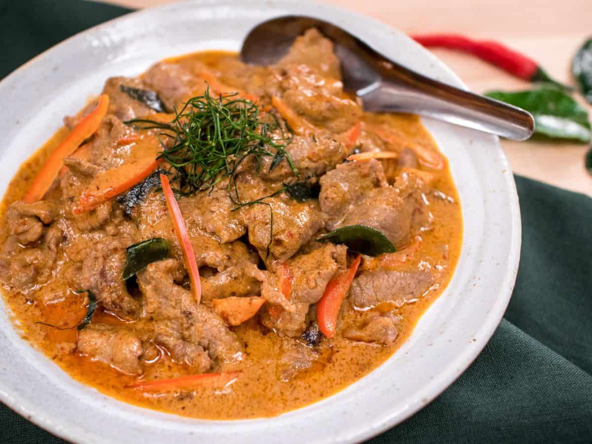 a bowl of panang beef curry with chilies and lime leaves in the background