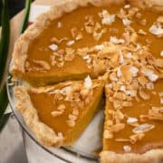 A whole pumpkin pie with one slice cut out with toasted coconut on top and pandan leaves on the side.