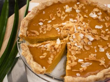 A whole pumpkin pie with one slice cut out with toasted coconut on top and pandan leaves on the side.