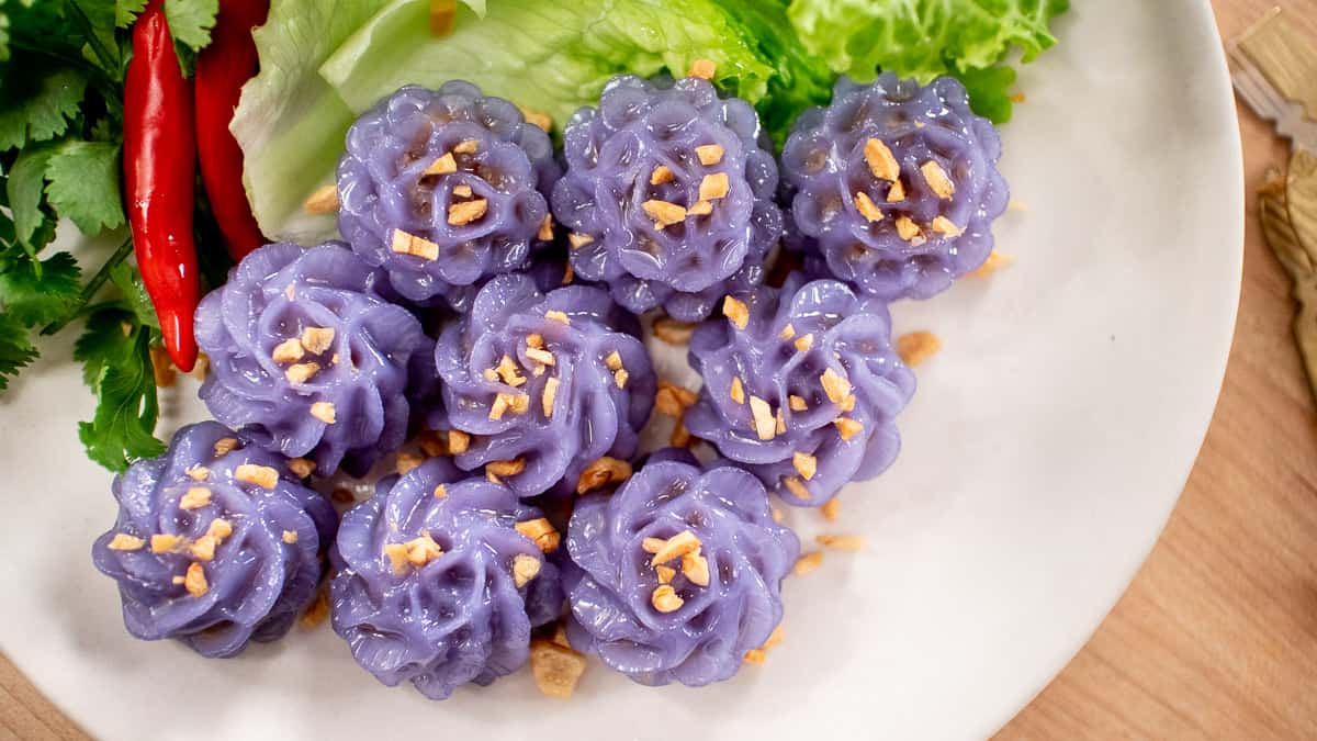 A plate of purple flower-shaped dumplings, with fried garlic on top, with a side of lettuce, chilies and cilantro.