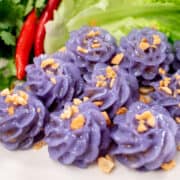 A plate of purple flower-shaped dumplings, with fried garlic on top, with a side of lettuce, chilies and cilantro.