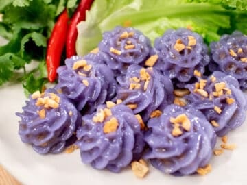 A plate of purple flower-shaped dumplings, with fried garlic on top, with a side of lettuce, chilies and cilantro.