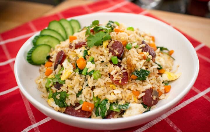 A bowl of chinese sausage fried rice with cucumber garnish on red tablecloth.