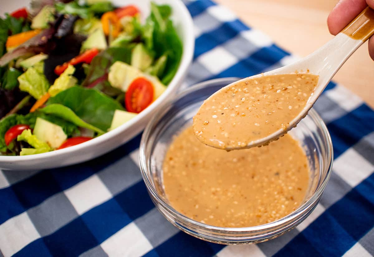 A spoonful of roasted sesame dressing being held over a bowl of dressing. With a green salad on the side. sp