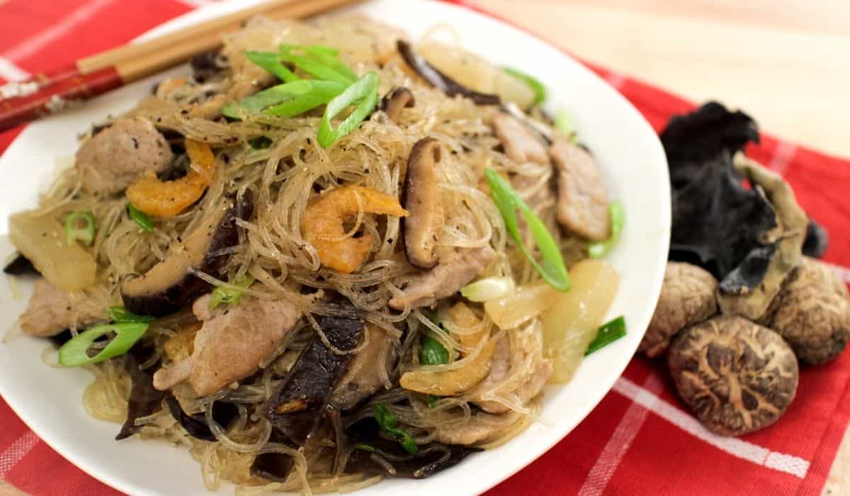 A bowl of glass noodle stir fry with dried shrimp, shiitake mushrooms and green onions on top.