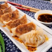 A plate of gyozas with crispy bottoms, with chili oil drizzled on them and dipping sauce on the side.