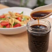 Stir fry sauce in a mason jar with a spoon pouring sauce into it. With a plate of a veggie stir fry in the background