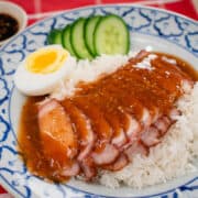 A plate of Chinese BBQ pork on rice with gravy on top. With a side of medium boiled egg and cucumber slices and a bowl of soy chili vinegar dippins sauce.