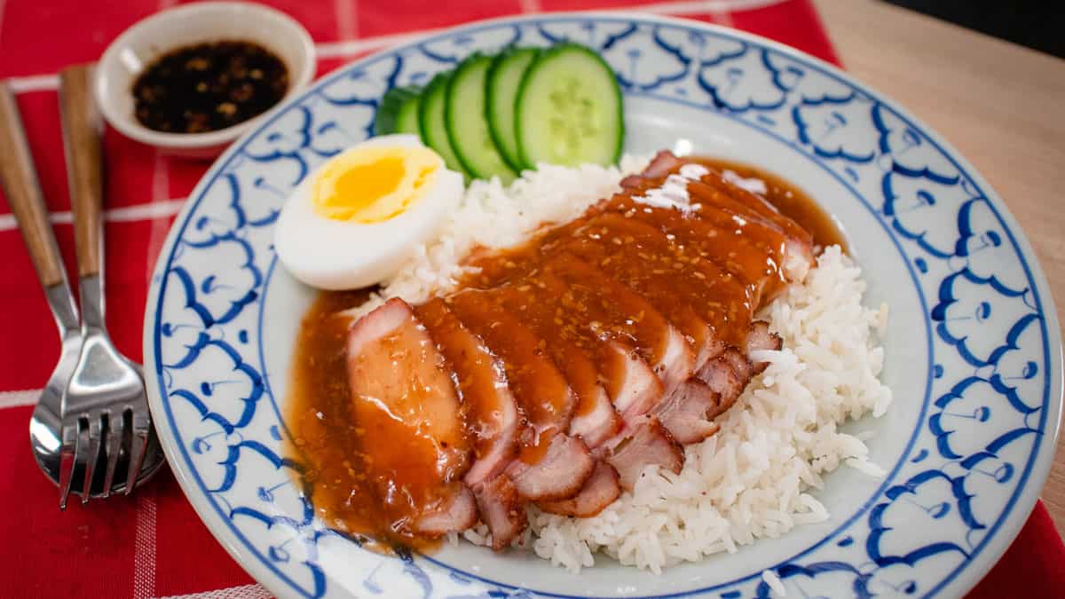 A plate of Chinese BBQ pork on rice with gravy on top. With a side of medium boiled egg and cucumber slices and a bowl of soy chili vinegar dippins sauce.