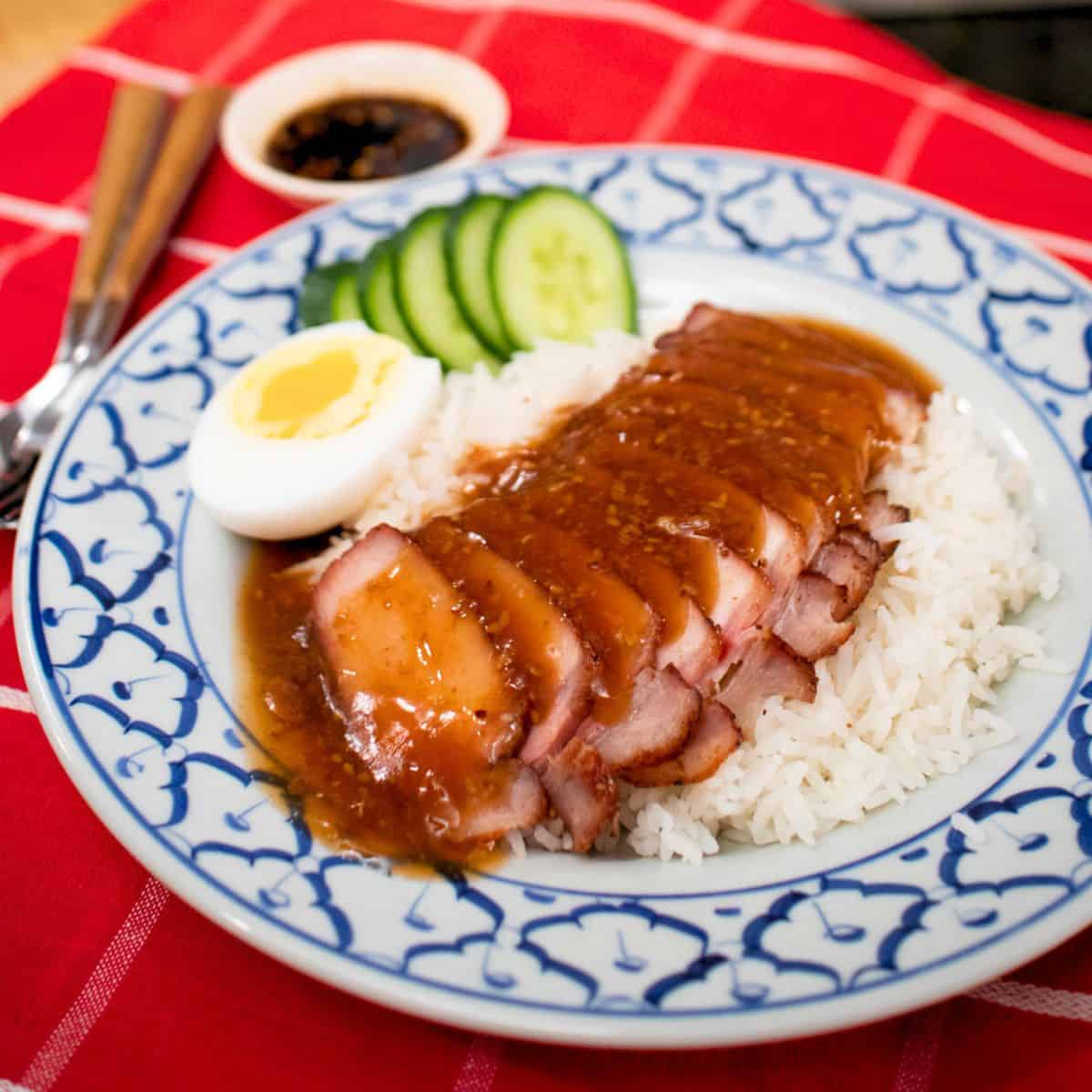 a plate of kao moo dang with a side of cucumber and boiled eggs and a bowl of dark soy sauce