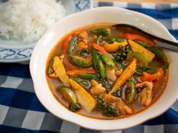 A bowl of jungle curry with baby corn, Thai eggplant, long beans, and chicken, with a plate of jasmine rice on the side.