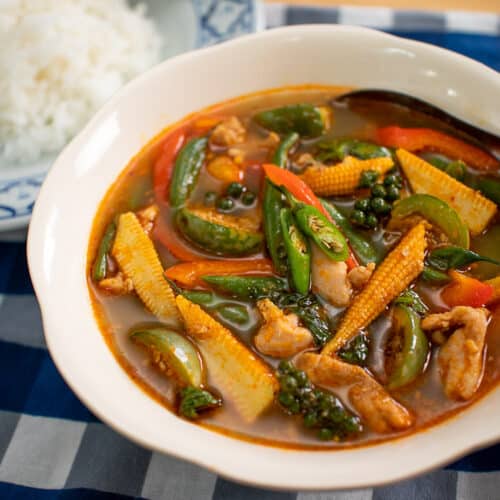 A bowl of jungle curry with baby corn, Thai eggplant, long beans, and chicken, with a plate of jasmine rice on the side.