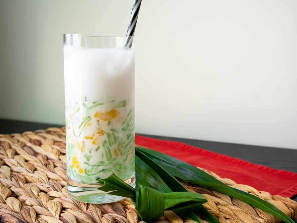 A glass of lodchong singapore in coconut milk with a straw in the glass and pandan leaves on the side.