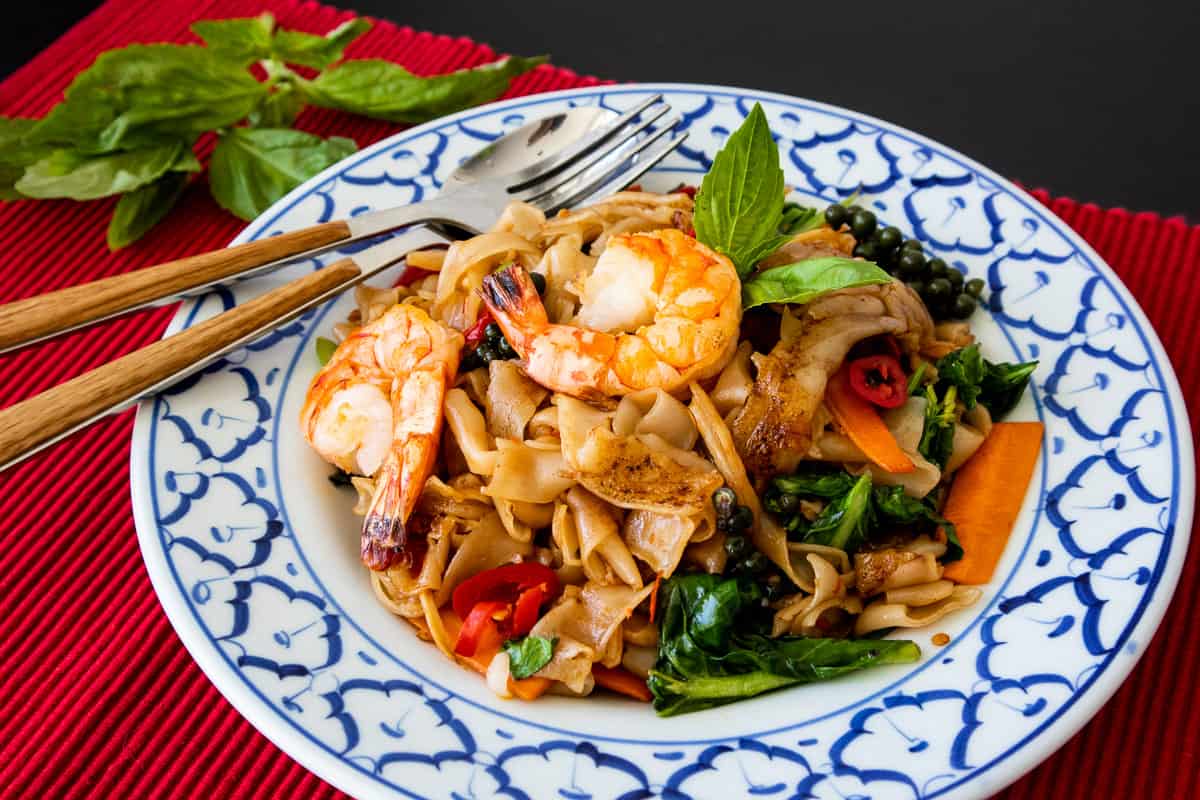 Pad kee mao is placed in a heaping pile on a white and blue ornate plate. The succulent shrimps are placed on the top left side of the pile while the vegetables are a beautiful contrast to the rich and flavourful noodles. A pair of chopsticks are placed on the left side. Herbs are placed on a red background beside the plate.