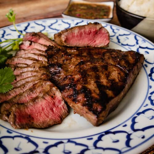 A plate of grilled steaks, some thinly sliced with dipping sauce and sticky rice on the side.