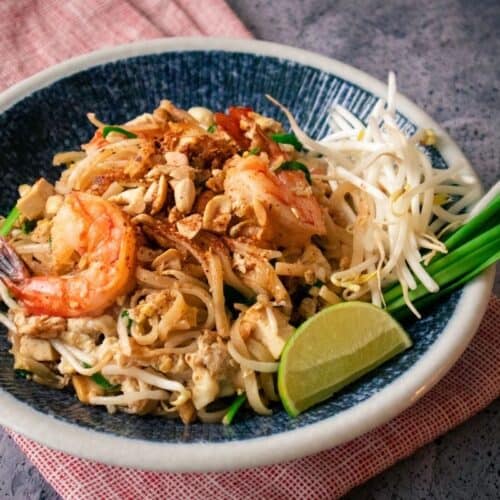 A plate of pad thai shrimp with lime and beansprouts and garlic chives on the side