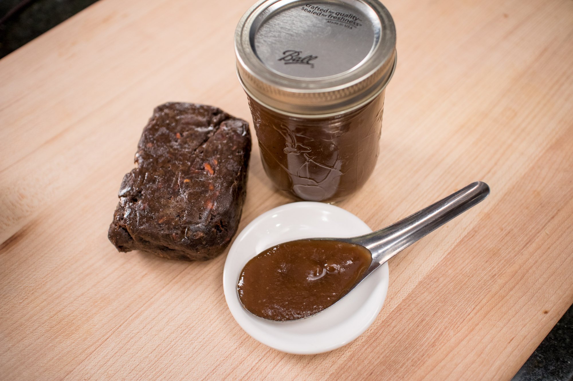 A block of tamarind pulp, a jar of tamarind paste, and a spoon filled with tamarind paste.