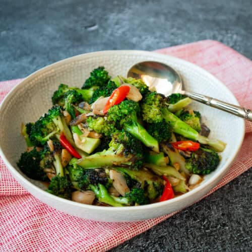 a plate of broccoli stir fry with salted fish, bits of chilies and a brass spoon and