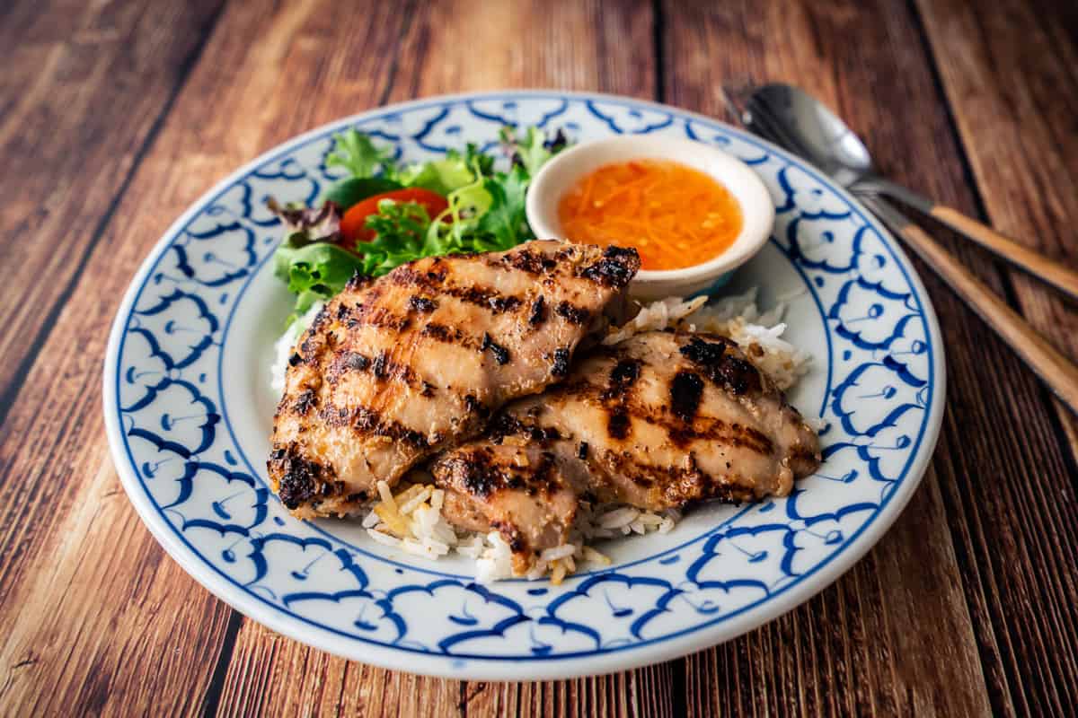A plate of grilled lemongrass chicken on rice with a side of nuac cham dipping sauce and salad greens.