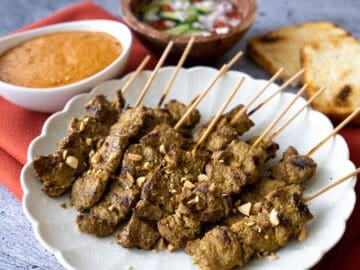 A plate of beef satay with a side of peanut sauce, pickles, and toast