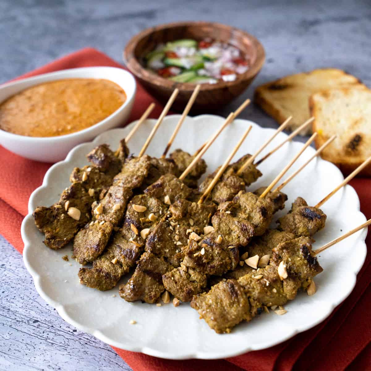 A plate of beef satay with a side of peanut sauce, pickles, and toast