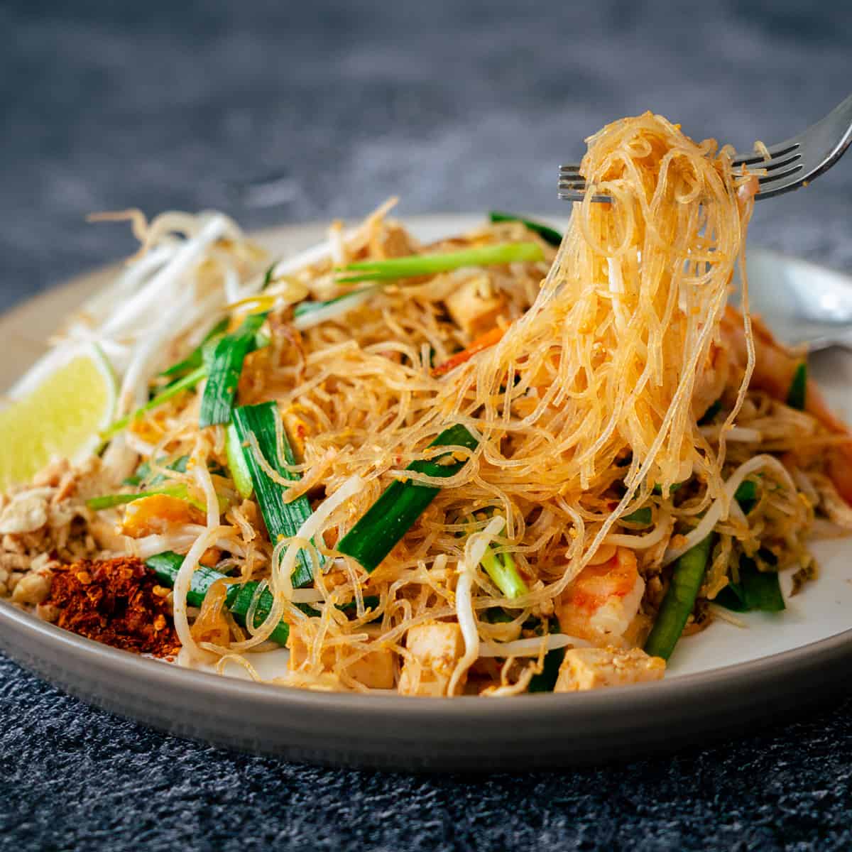 A fork full of golden glass noodles are being pulled out of a pile of pad thai woonsen. Its vegetables and ingredients glow and glisten in contrast to the refreshing garnishes of lime and beansprouts. The chili powder and peanuts give a wonderful splash of colour.