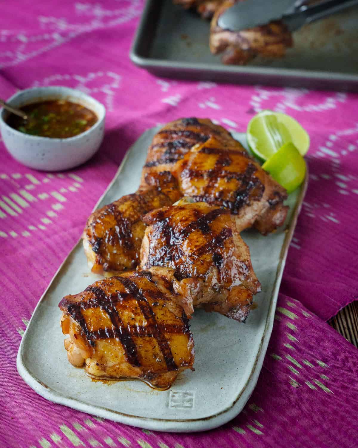 a plate of grilled chicken thighs with lime wedges on the side and a bowl of dipping sauce.