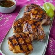 a plate of grilled chicken thighs with lime wedges on the side and a bowl of dipping sauce.