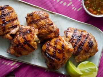 a plate of grilled chicken thighs with lime wedges on the side and a bowl of dipping sauce.