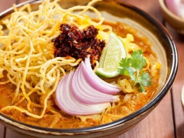 a bowl of khao soi with shallots, lime, cilantro and fried chilies on top