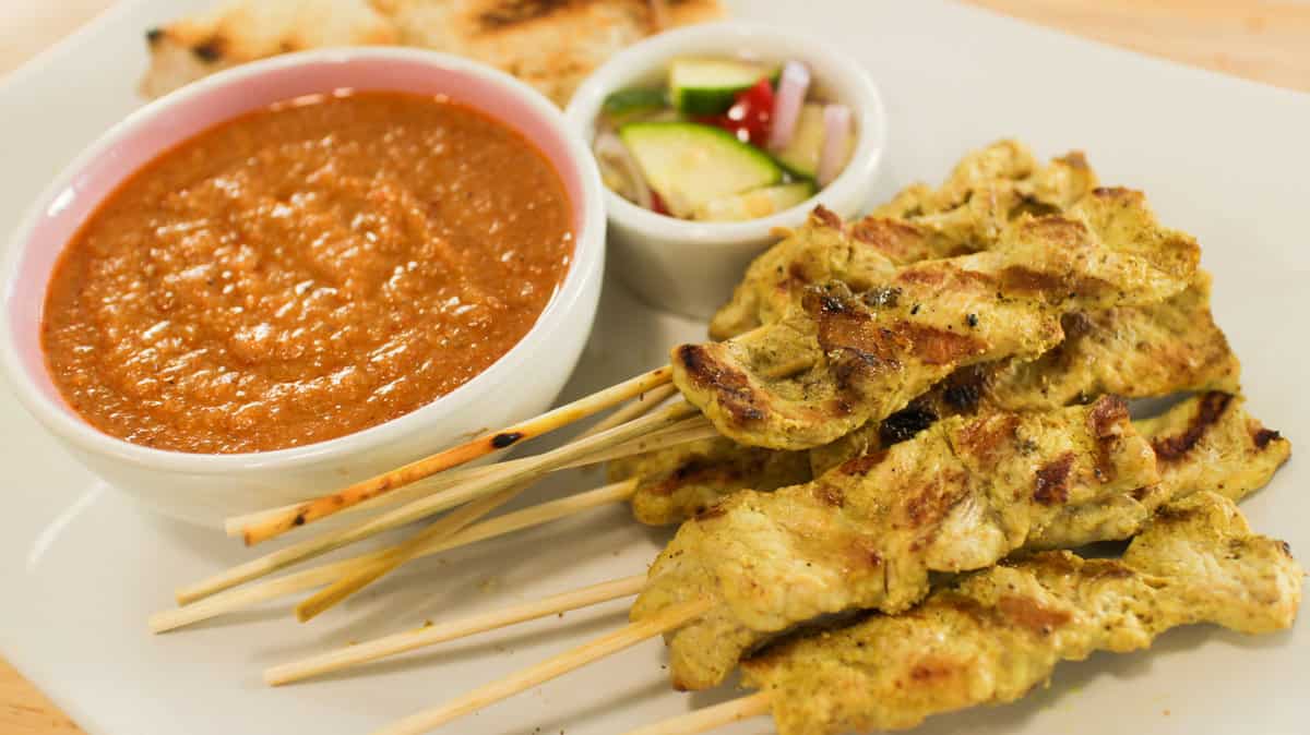 Skewers of pork Satay perfectly grilled and placed in a uniform pile on a white plate. It is paired with a rich peanut sauce and veggie side.