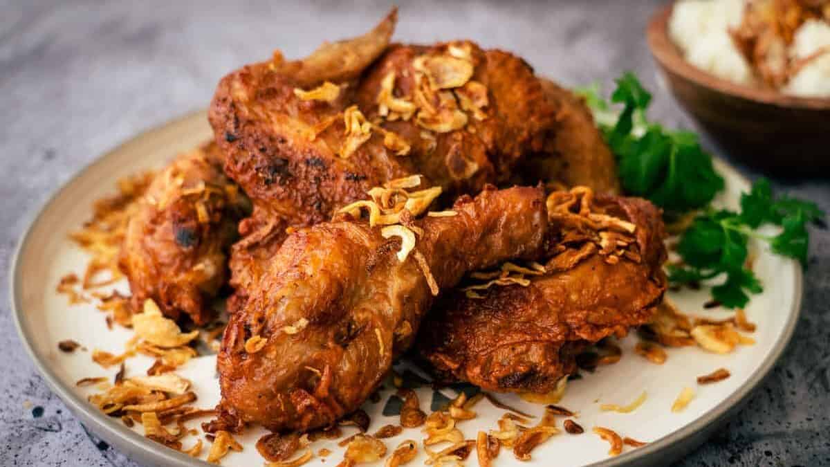 Golden brown Hat Yai fried chicken wings are placed in a delicious pile on a ceramic plat with crispy fried onions and garnished with a sprig of herbs. It is placed on a grey stone background.