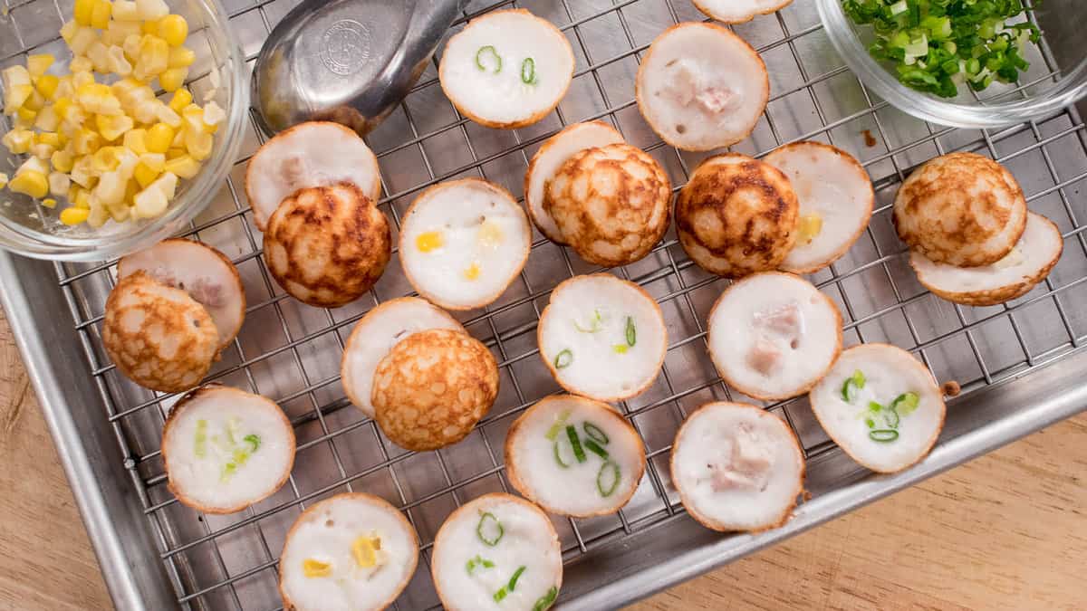 Various kanom krok halves are placed cut open side up or down on a wire rack. The fillings of corn, taro cubes, or spring onions peak through the cooked batter. The are two clear bowls filled with corn or spring onions.