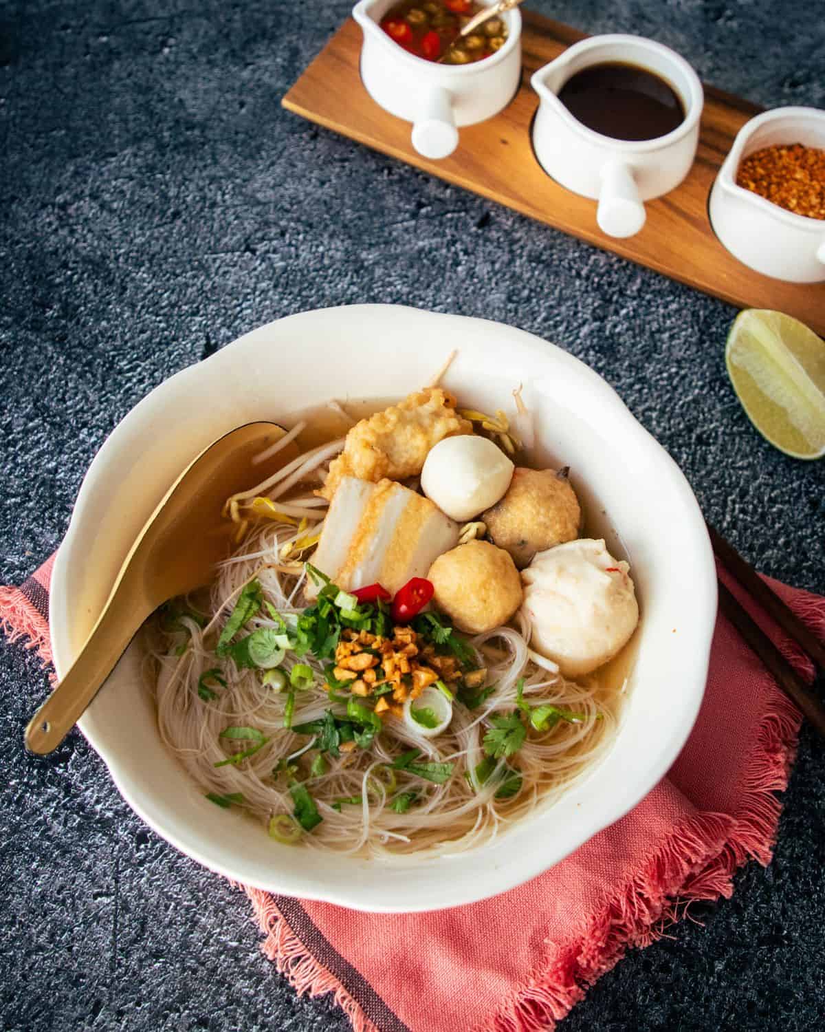 A bowl of noodle soup with fish cakes with a lime wedge and condiments on the side