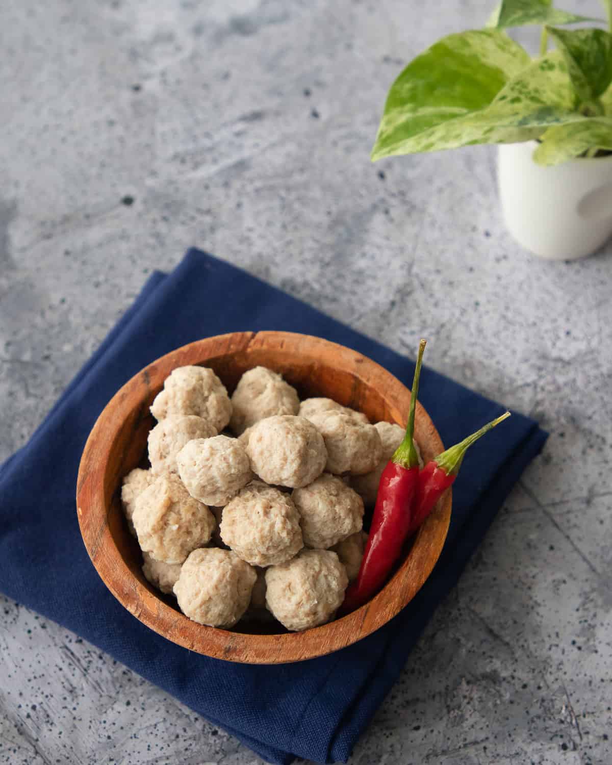 a bowl of moo deng meatballs with 2 chilies and a plant in the background