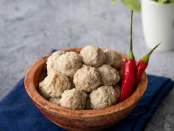 a bowl of moo deng meatballs with 2 chilies and a plant in the background