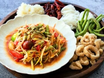 papaya salad lao style on a plate with a side of crispy pork rind, long beans, rice noodles, dried chilies and sticky rice