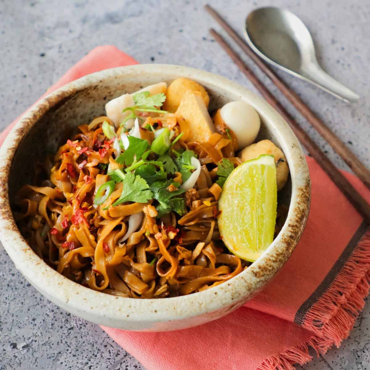 A bowl of tossed noodles with chopsticks and spoon on the side.