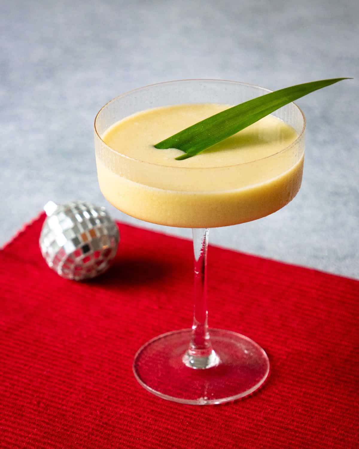 mango sticky rice cocktail in a coupe glass with a pandan leaf garnish and a small disco ball in the background.