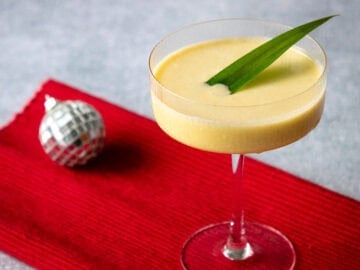 mango sticky rice cocktail in a coupe glass with a pandan leaf garnish and a small disco ball in the background.